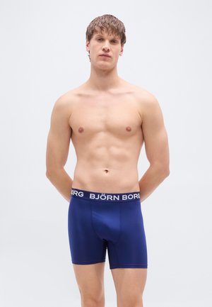 Caleçons boxers bleu marine en tissu lisse, arborant une ceinture blanche avec logo et une coupe ajustée offrant une couverture modérée.