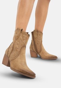 Botas de tobillo de cuero marrón con punta afilada, costuras decorativas, lengüetas de tirón y tacones en bloque. Textura suave con patrones destacados.