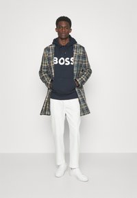 Cappotto a quadri verde e blu sopra un hoodie blu navy con la scritta bianca "BOSS", abbinato a pantaloni bianchi e sneakers bianche.