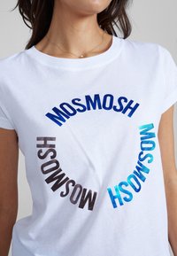 Vitt bomullströja med cirkulärt textmönster i olika nyanser: blått, turkost och brunt tryckt över framsidan. Kort ärm.