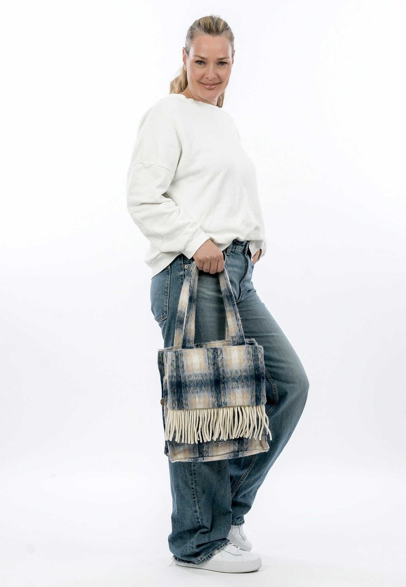 Blaue und beige karierte Handtasche mit Fransen, aus weichem Stoff gefertigt. Zwei robuste Tragegriffe für einen einfachen Transport. Lässiger Stil, kombiniert mit einem Jeans-Outfit.