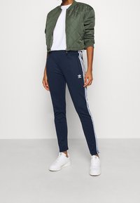 Grön quiltad bomberjacka, vit t-shirt, marinblå sweatpants med vita ränder och vita sneakers. Har en logotyp på byxorna.