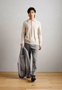 Pulover tricotat bej deschis cu guler, pantaloni gri croiți și un sacou gri ținut într-o mână. Pantofi întunecati completează ținuta.