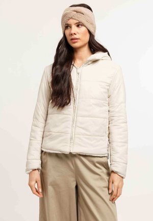 Woman with long dark hair wearing a beige quilted jacket, matching wide-leg pants, and a fuzzy headband, standing against a plain background.