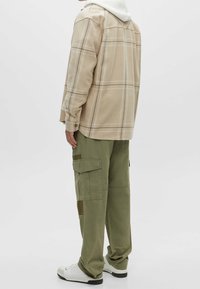 Personne portant une veste à carreaux beige, un pantalon cargo vert olive, des baskets blanches, debout contre un fond clair uni, vue de profil.