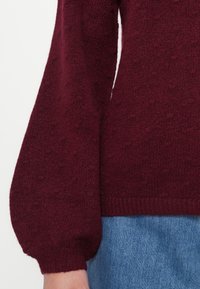 Burgundy kötött pulóver texturált kivitelben, puffos ujjal és bordázott aljával, farmer szoknyával párosítva.
