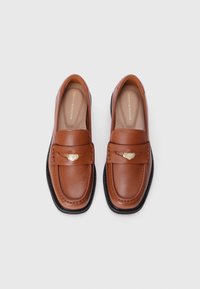 Mocassins en cuir marron avec une texture lisse, un accent doré et une semelle en caoutchouc noir contrastante. Design slip-on classique avec un style discret.