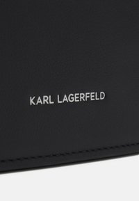 Svart lädermaterial med en texturerad yta. Har silverprägling med "KARL LAGERFELD"-logotyp och noggrann sömnad längs kanten.