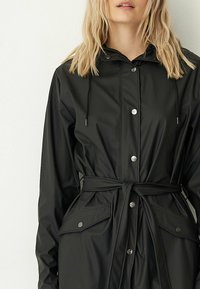 Zwarte waterdichte jas met een capuchon, drukknoopjes, een strakke taille met een strikceintuur en zijzakken. Soepele textuur met een matte afwerking.