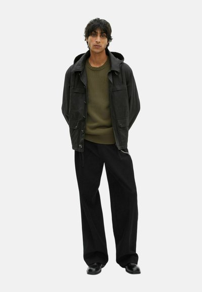 Schwarze Kapuzenjacke über einem olivgrünen Pullover, kombiniert mit schwarzen weitem Hosen und schwarzen Schuhen. Die Jacke hat sichtbare Taschen.