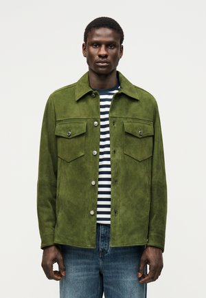 Muž v zelenej semišovej bunde cez čierno-biele pruhované tričko a modré džínsy, stojaci pred jednoduchým svetlým pozadím.