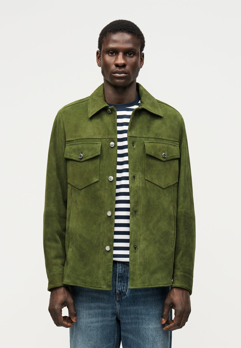 Άνδρας με πράσινο σουέντ σακάκι πάνω από μαύρο και άσπρο ριγωτό πουκάμισο και μπλε τζιν, στέκεται μπροστά σε απλό φωτεινό φόντο.