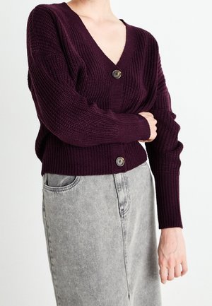 Cardigan en tricot violet foncé à col en V, manches longues, avec une fermeture centrale à boutons et une texture côtelée, associé à une jupe en denim gris.