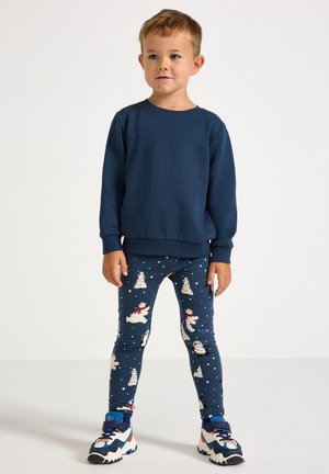 Marineblå sweatshirt med ribbede manchetter, parret med mønstrede leggins med snemænd og træer mod en mørkeblå baggrund. Farverige sneakers.