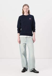 Sudadera azul marino con cuello redondo y logo bordado en el pecho, combinada con jeans de pierna ancha azul claro y zapatos negros.