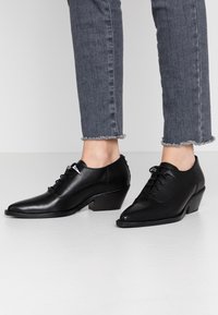 Scarpe in pelle nera con punte appuntite e tacco basso a blocco. Presentano una chiusura con lacci, texture liscia e un sottile dettaglio di cucitura.