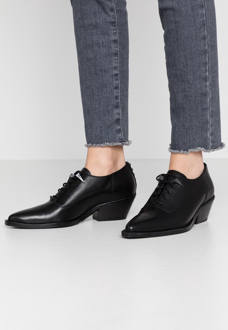 Scarpe in pelle nera con punte appuntite e tacco basso a blocco. Presentano una chiusura con lacci, texture liscia e un sottile dettaglio di cucitura.