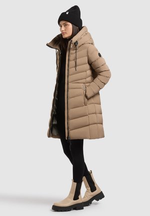 Beige gewatteerde jas met een hoge kraag, verticale stiksels en ritssluiting. Gedragen over zwarte legging en beige enkellaarsjes met zwarte accenten.