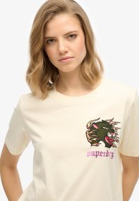 T-shirt crème à col rond, ornée d'un motif détaillé de tête de panthère verte brodée et du mot "superdry" en rose.