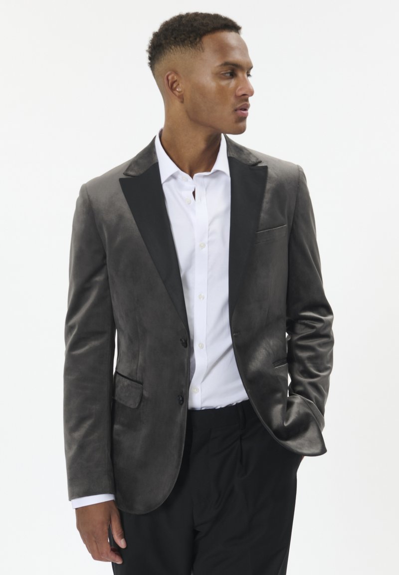Mand iført en mørkegrå fløjl blazer med sorte klapper, hvid skjorte og sorte bukser, ser til venstre med den ene hånd i lommen.