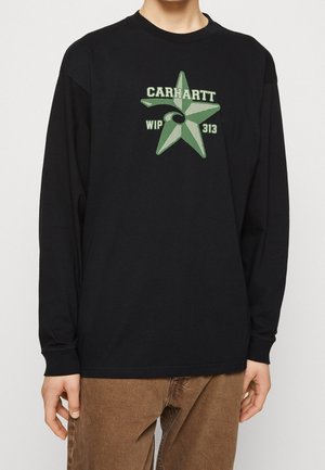 Sort langærmet skjorte med grøn stjerne og teksten "CARHARTT WIP 313" båret af person med brune bukser mod hvid baggrund.
