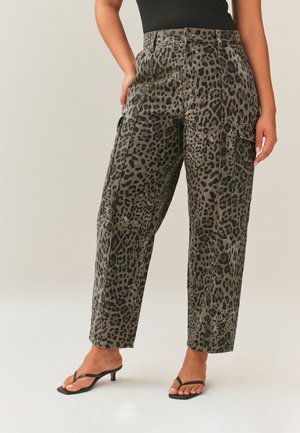 Femme portant un pantalon cargo taille haute à jambes larges avec un imprimé léopard gris et noir et des sandales à talons noires, debout devant un fond uni.