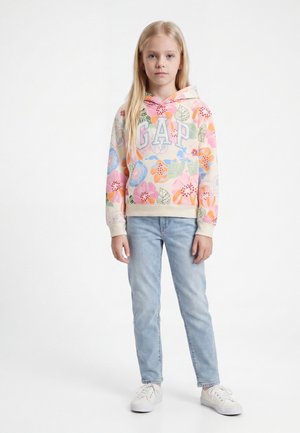 Ένα νεαρό κορίτσι στέκεται απέναντι, φοράει ανοιχτό μπλε τζιν, λευκά αθλητικά παπούτσια και παστέλ φλοράλ φούτερ με το γράμμα «GAP».