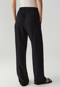 Zwarte, lichte, wijde broek met een elastische tailleband en zijzakken, van achteren getoond, rustend op een neutrale achtergrond.