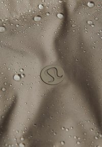 Burgundska tkanina s kapljicama vode, s istaknutim kružnim logom u donjem desnom kutu. Tekstura izgleda glatko i lagano odražava svjetlost.