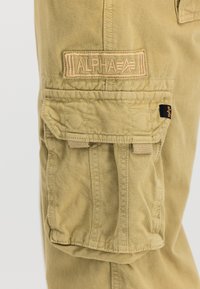 Khaki Cargo-Hosentasche mit Klappe, Nahtdetails und beigem Stoffpatch mit der Aufschrift "ALPHA" über der Tasche am linken Bein.