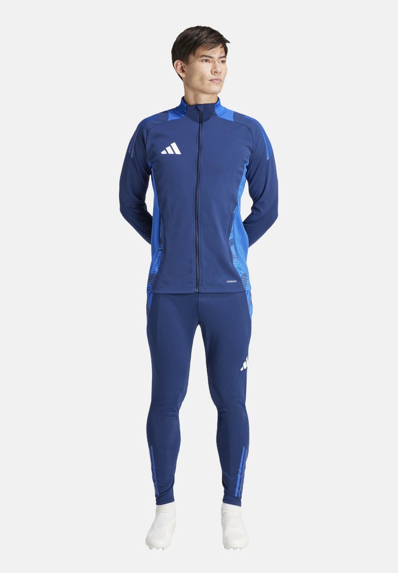 Tuta sportivo di colore blu scuro con accenti blu chiaro, giacca con zip e pantaloni aderenti, realizzati in tessuto liscio ed elasticizzato. Logo Adidas visibile.