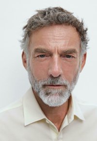 Primo piano di un uomo anziano con capelli e barba grigi, che indossa una camicia beige chiaro, su uno sfondo bianco semplice. I tratti del viso sono ben visibili.
