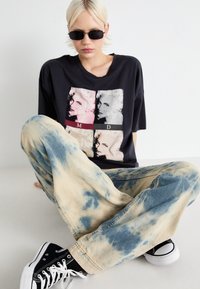 Camiseta negra de gran tamaño con un diseño gráfico en rosa y gris, combinada con jeans holgados en lavado ácido azul y beige, y zapatillas negras.