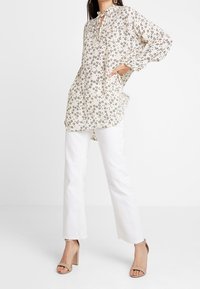 Kukkakuvioinen, kevyt beige pusero sidotulla kaula-aukolla; yhdistetty valkoisiin suoriin housuihin ja nude-värisiin paksupohjaisiin sandaaleihin.