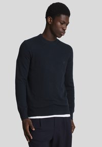 Suéter de cuello redondo en un oscuro tono navy, con puños y dobladillo acanalados, que presenta un pequeño logo en el pecho. Textura de punto suave.