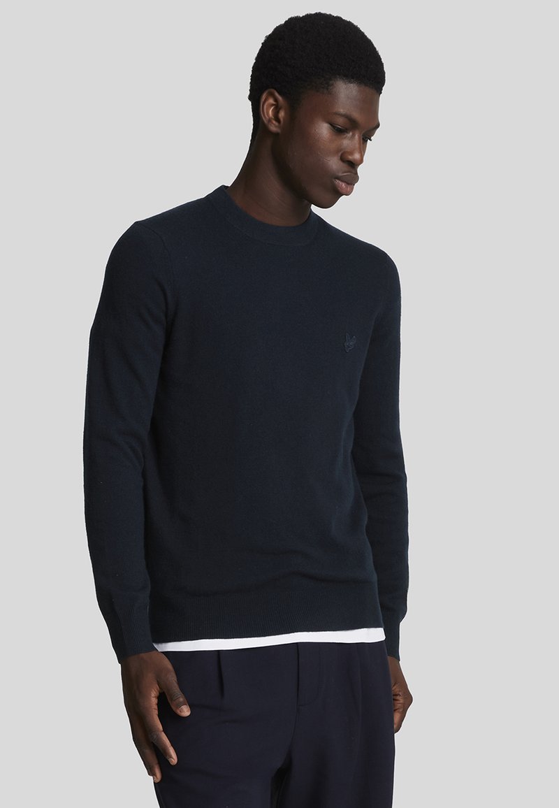 Suéter de cuello redondo en un oscuro tono navy, con puños y dobladillo acanalados, que presenta un pequeño logo en el pecho. Textura de punto suave.