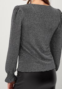 Schwarzes, schimmerndes Oberteil mit langen Ärmeln, gepufftem Schulterdetail und gewelltem Saum. Es hat eine glatte Textur und eine figurbetonte Silhouette.