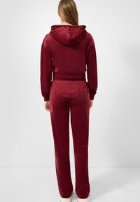 Vrouw gezien van achteren in een kastanjebruine velours hoodie en bijpassende broek met reliëf "Von Dutch" logo's op de achterkant.
