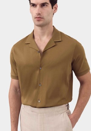 Mann trägt braunes Kurzarmhemd mit Knöpfen, in hellbeige, hochtaillierte Hose gesteckt, Hand in der Tasche.