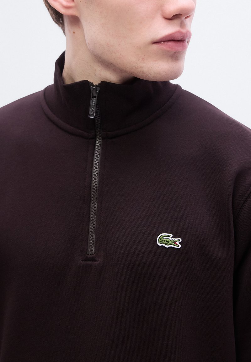 Pull à demi-fermeture éclair de couleur brun foncé en matière douce, avec un petit logo crocodile brodé vert et blanc sur la poitrine.