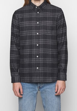 Mand iført mørkegrå og sortternet langærmet skjorte med knapper samt lyseblå jeans, stående mod en ensfarvet baggrund.