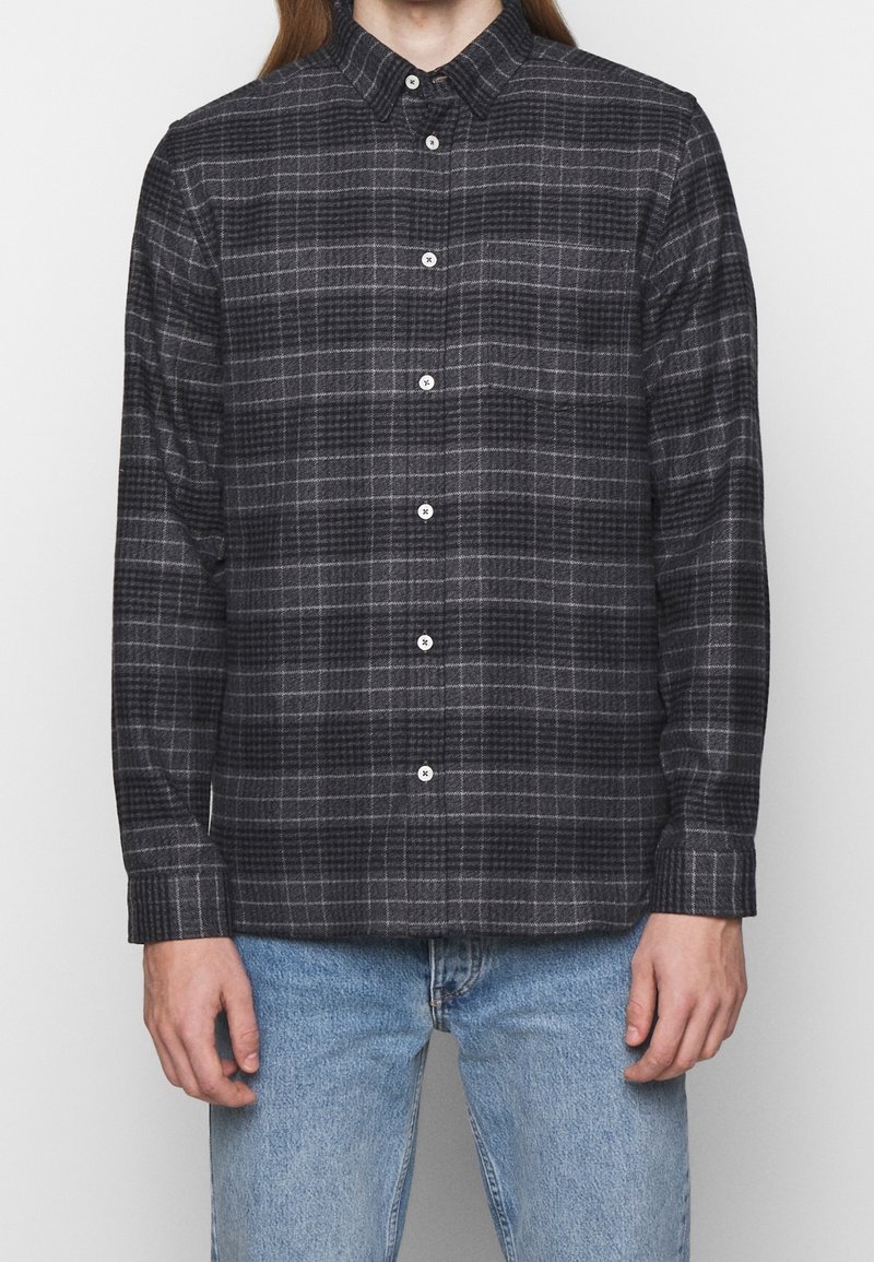 Homme portant une chemise à manches longues à carreaux gris foncé et noir avec un jean bleu clair, debout devant un fond uni.