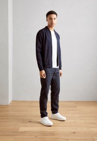 Navy cardigan over a white t-shirt, paired with dark pants and white sneakers. Smooth fabric and straight-fit design, standing on wooden floor.