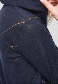 Marinblå stickad tröja med huva, texturerad tyg och gyllene accentstickningar i diagonala linjer över armarna och axlarna.