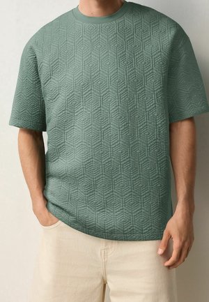 Homme portant une chemise courte matelassée verte avec des motifs géométriques et un pantalon beige clair, une main dans la poche.