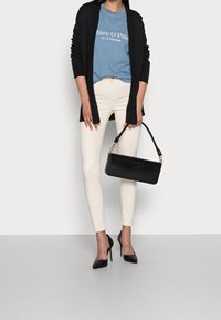 Cardigan lungo nero sopra una t-shirt blu con testo bianco, jeans skinny crema e tacchi a punta neri. Tiene una borsa nera con dettagli dorati.