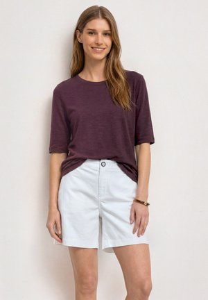 Jeune femme aux longs cheveux portant un t-shirt violet à manches courtes et un short blanc, debout contre un mur clair, souriante.