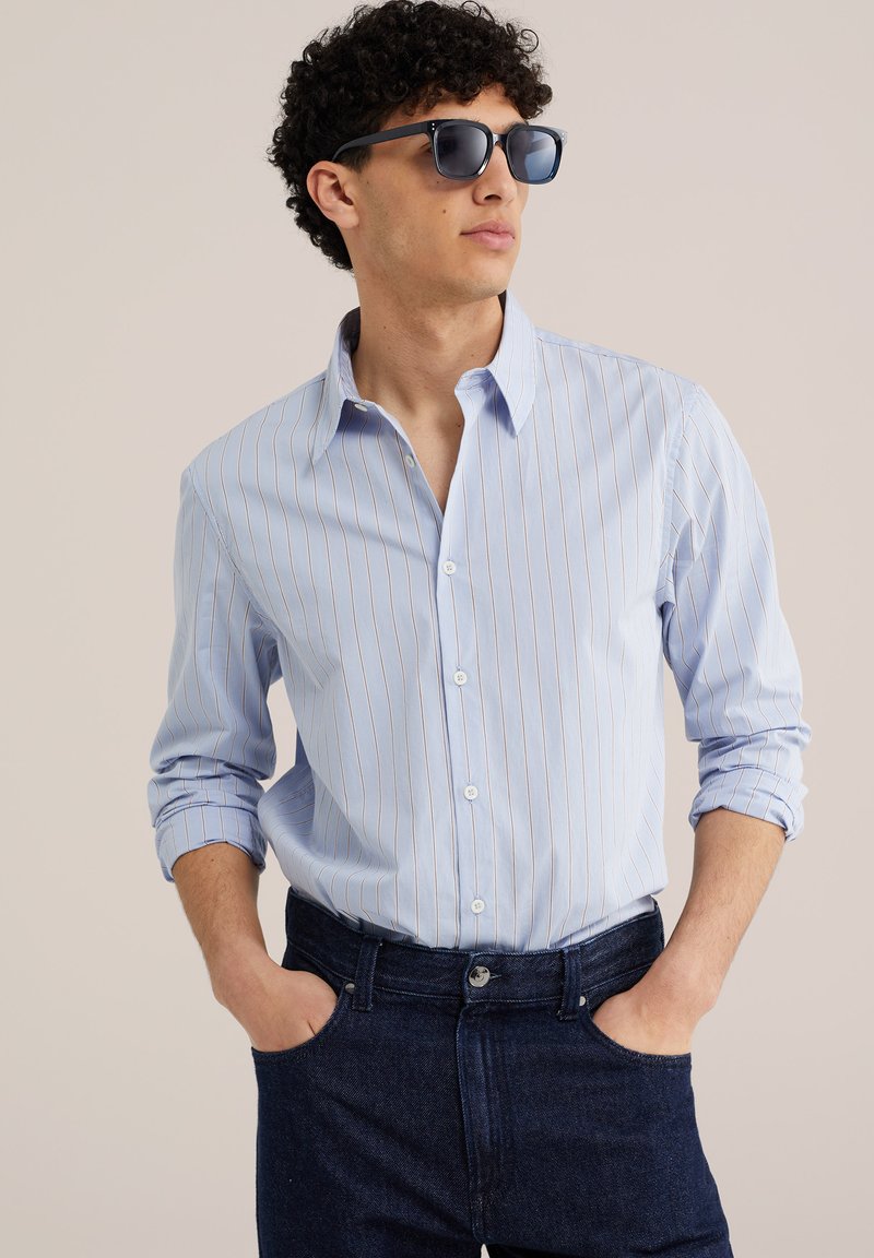 Homme portant une chemise bleue à fines rayures avec manches retroussées, jean foncé et lunettes de soleil noires, mains dans les poches, regardant sur le côté.