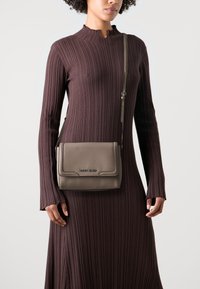 Abito lungo a maniche lunghe marrone a coste con collo alto, abbinato a una borsa in pelle grigia con patta e dettagli in metallo.