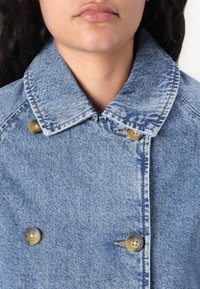 Denim jakk, millel on terav krae, traditsiooniline kahekordne disain, helesinine värv ja kaks messingi nuppu krae lähedal.
